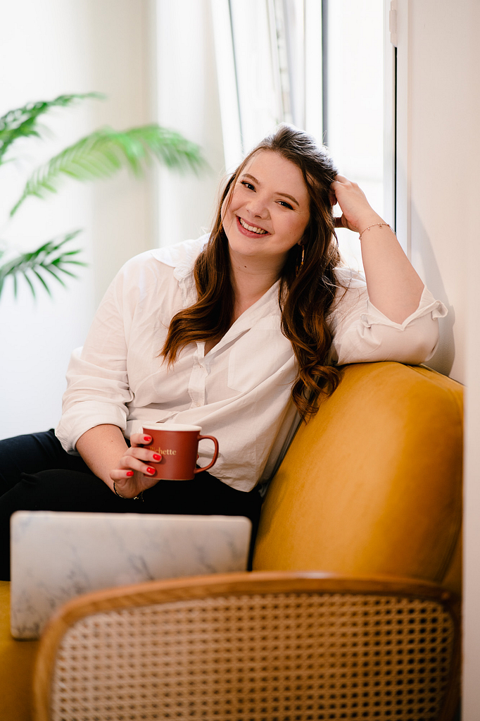 Une femme entrepreneure regarde l'objectif en souriant, une tasse de café à la main et assise sur un canapé jaune.
