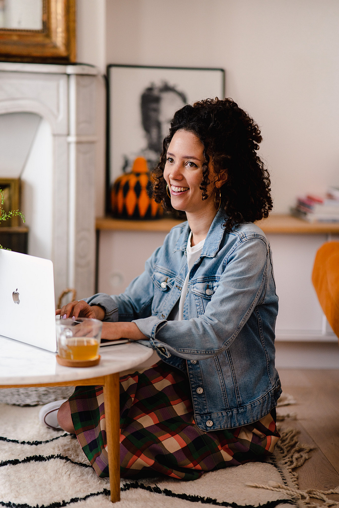 Une femme, entrepreneure, assise par terre devant la table basse en train de travailler sur son PC avec un grand sourire.
